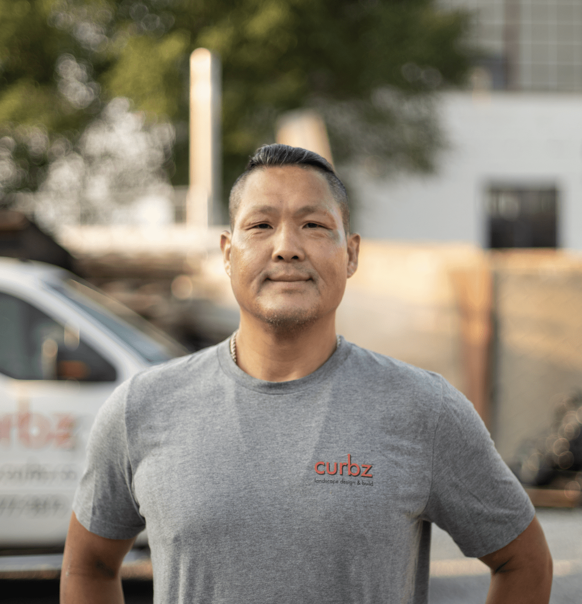 A person stands confidently outdoors in a gray "curbz" t-shirt, with trees and a vehicle featuring blurred text in the background.